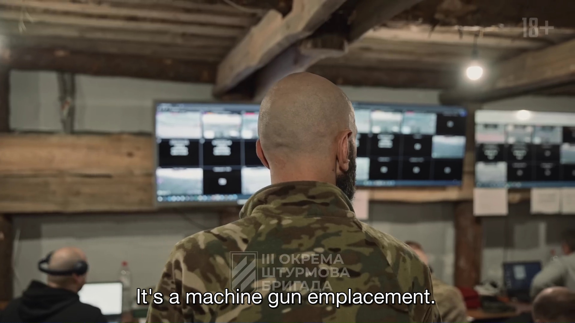 Un homme de dos, crâne rasé, barbe et treillis, devant plusieurs écran, sous un plafond bas fait de poutres apparentes.