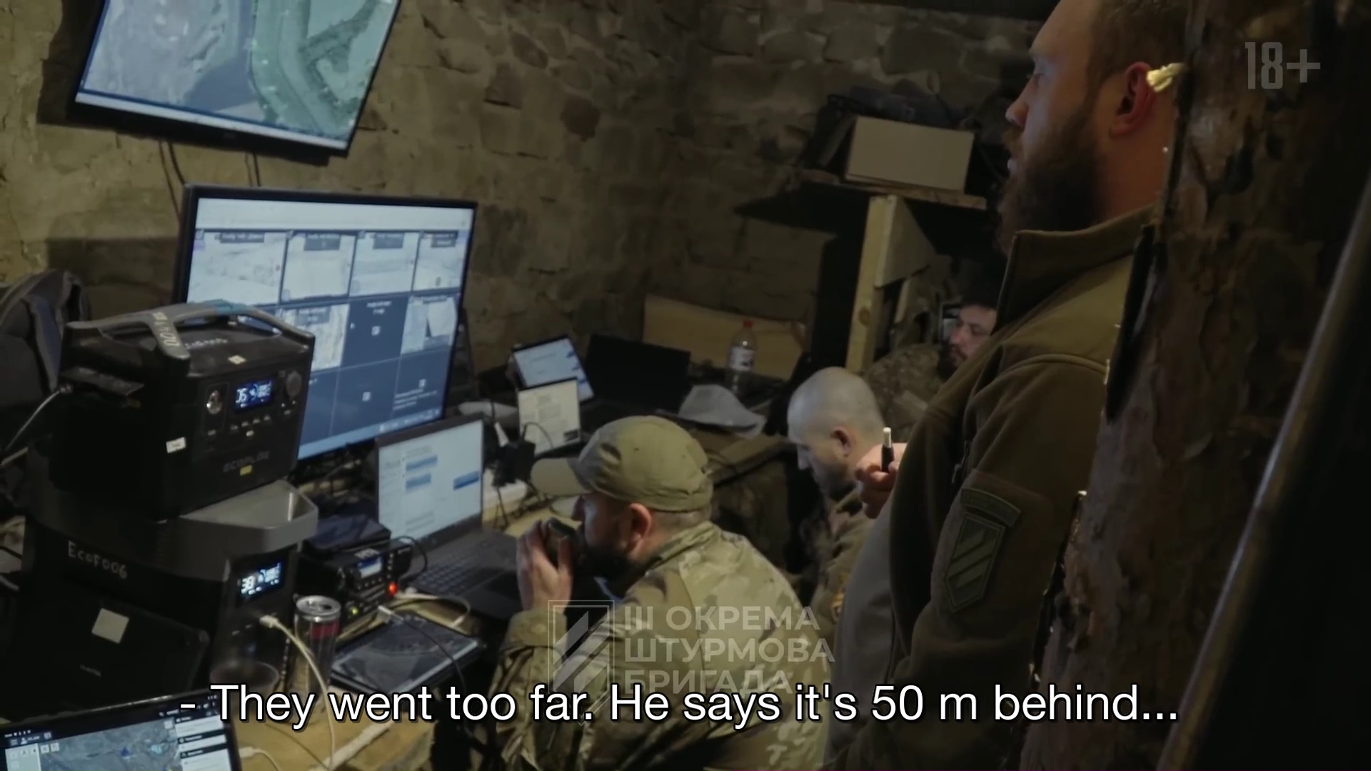 Un homme debout et trois assis devant des écrans, en treillis, dans une pièce sombre. En sous-titre : "They went too far. He says it's 50 m behind..."