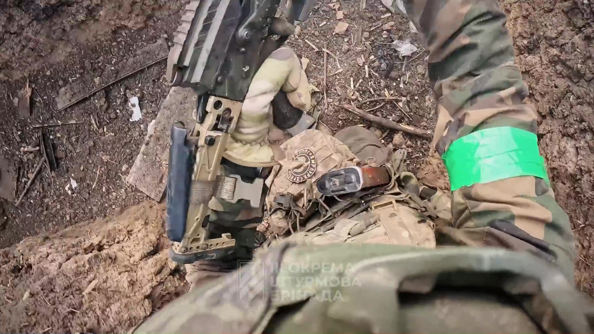 Vue subjective vers le sol. Des mains gantées en treillis tiennent une mitrailleuse. Le sol est terreux, comme dans une tranchée.