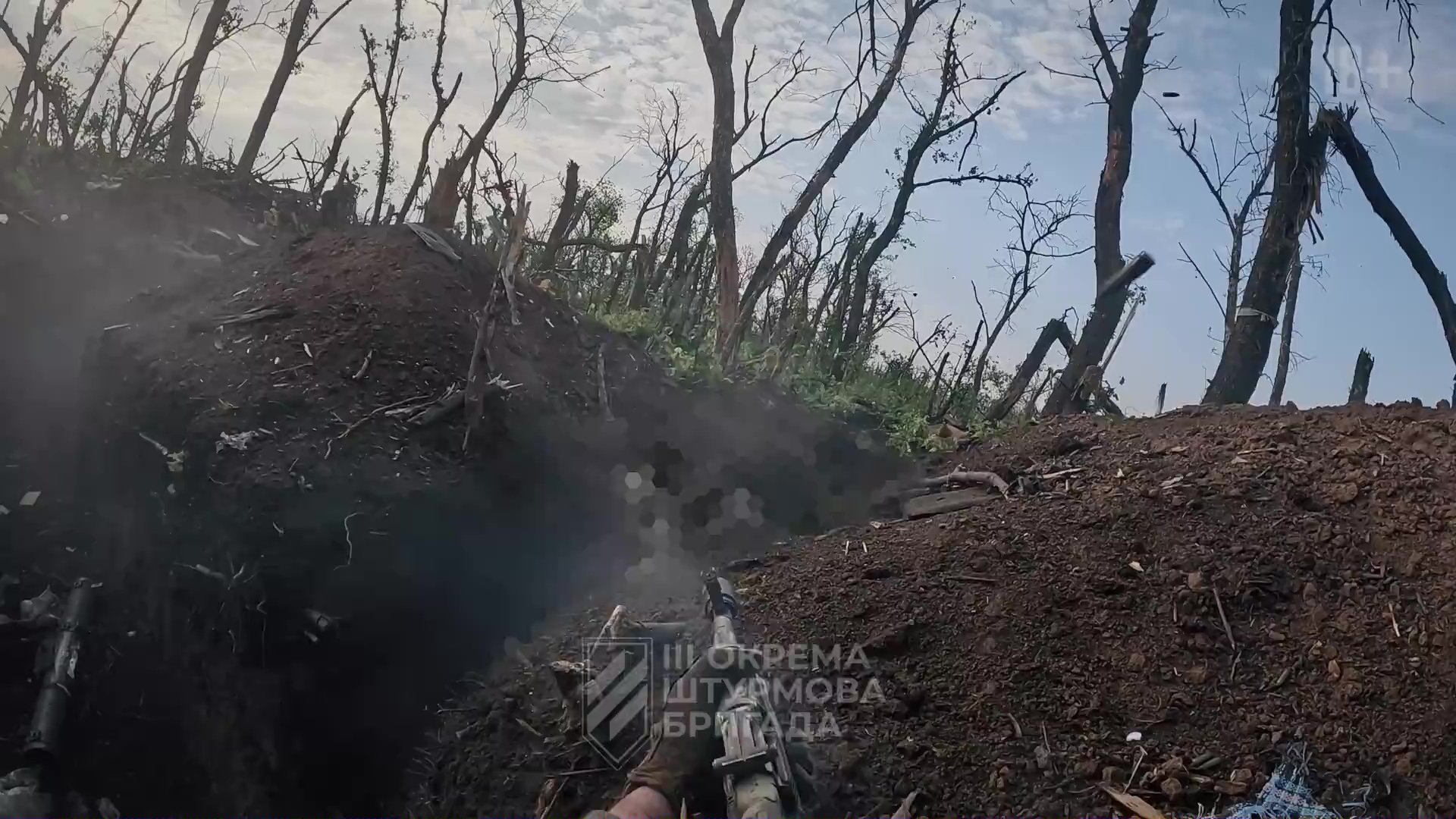 Vue subjective d'un tir de mitrailleuse depuis une tranchée. Le ciel est bleu, les arbres sont tous abîmés et brûlés, il n'en reste que des troncs rachitiques.