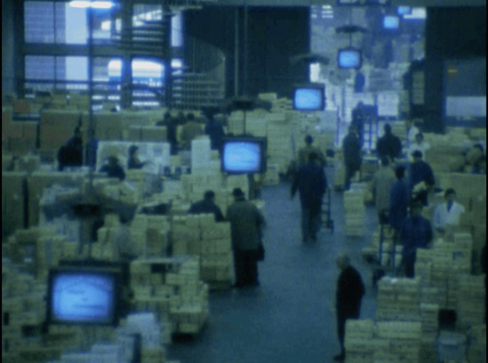 Vue vidéo, peu définie. Une allée avec, de part et d'autre, des palettes remplies et, à intervalles réguliers, des écrans cathodiques.