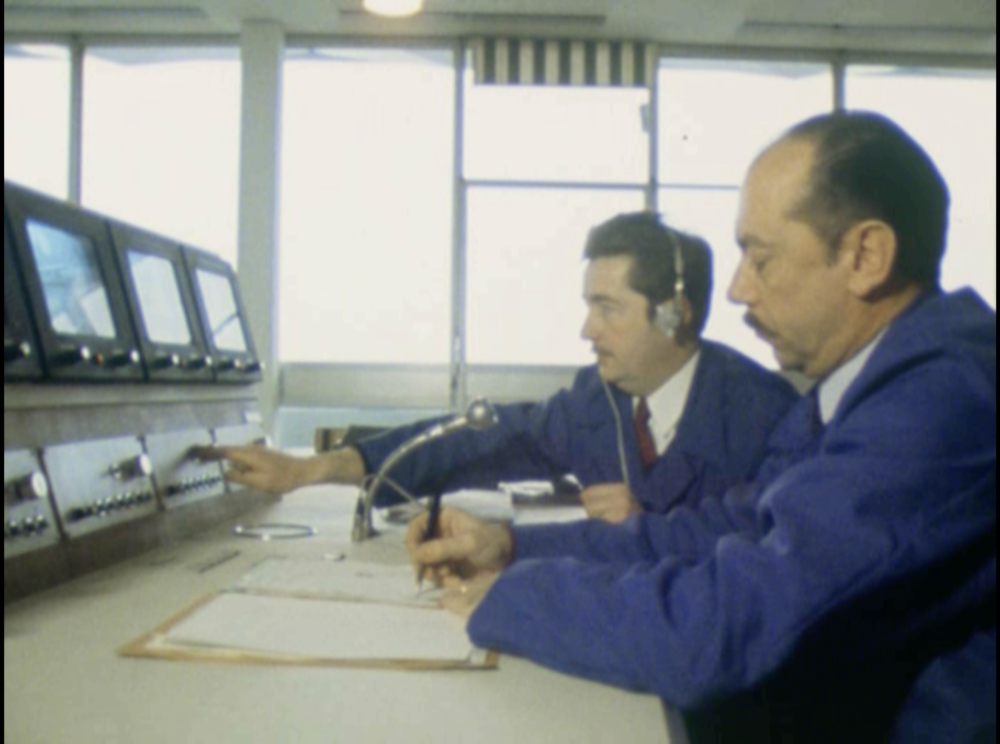 Des opérateurs en bleu de travail, prennent des notes face à des écrans.