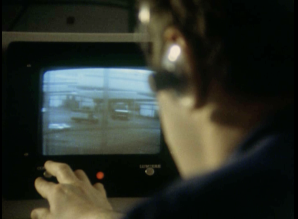 À l'avant-plan, flou, un opérateur de trois-quarts dos avec un casque sur les oreilles. Il touche un potentiomètre sous un petit écran cathodique qui affiche des camions.