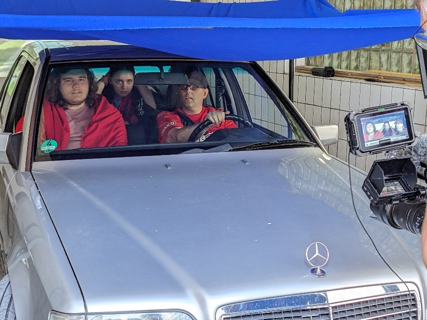 Trois jeunes gens sont assis dans une voiture. Ils portent des tricots et des écharpes aux couleurs du club de foot local. Le conducteur porte une casquette et des lunettes. Ses yeux sont rivés sur le rétroviseur intérieur. A droite, on aperçoit une caméra en train de filmer la scène. La voiture, qu’on voit de face, est recouverte d’une toile bleue et garée dans un espace carrelé de blanc.  