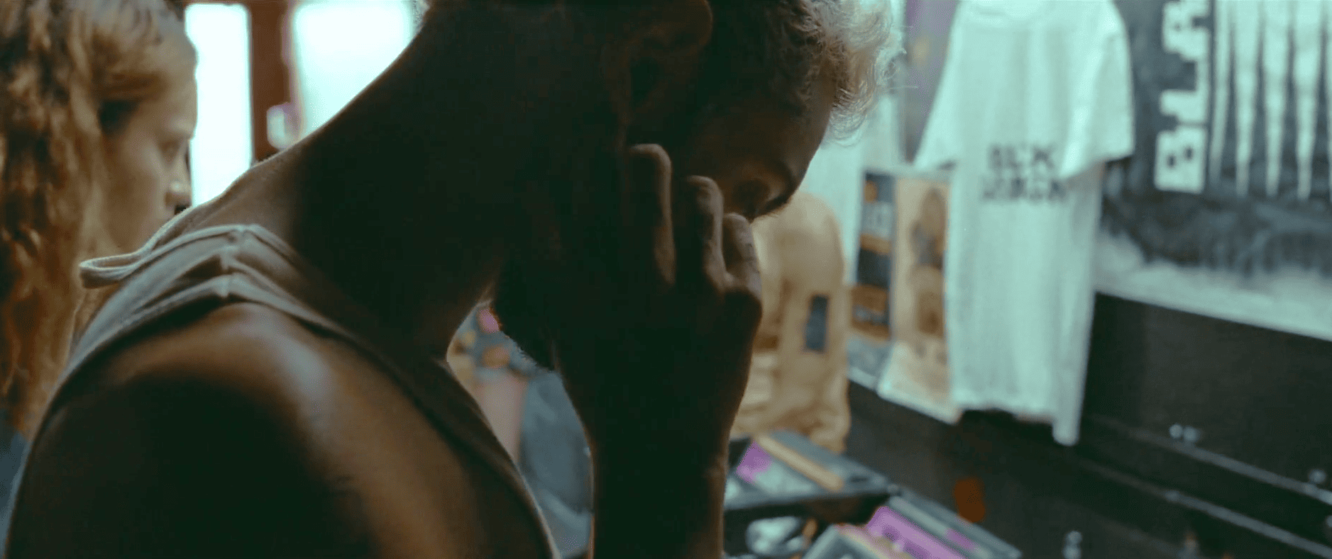 Un jeune homme de profil, en marcel blanc, baisse légèrement la tête et se touche l'oreille droite de l'index droit, penché au-dessus de bac de vinyls.