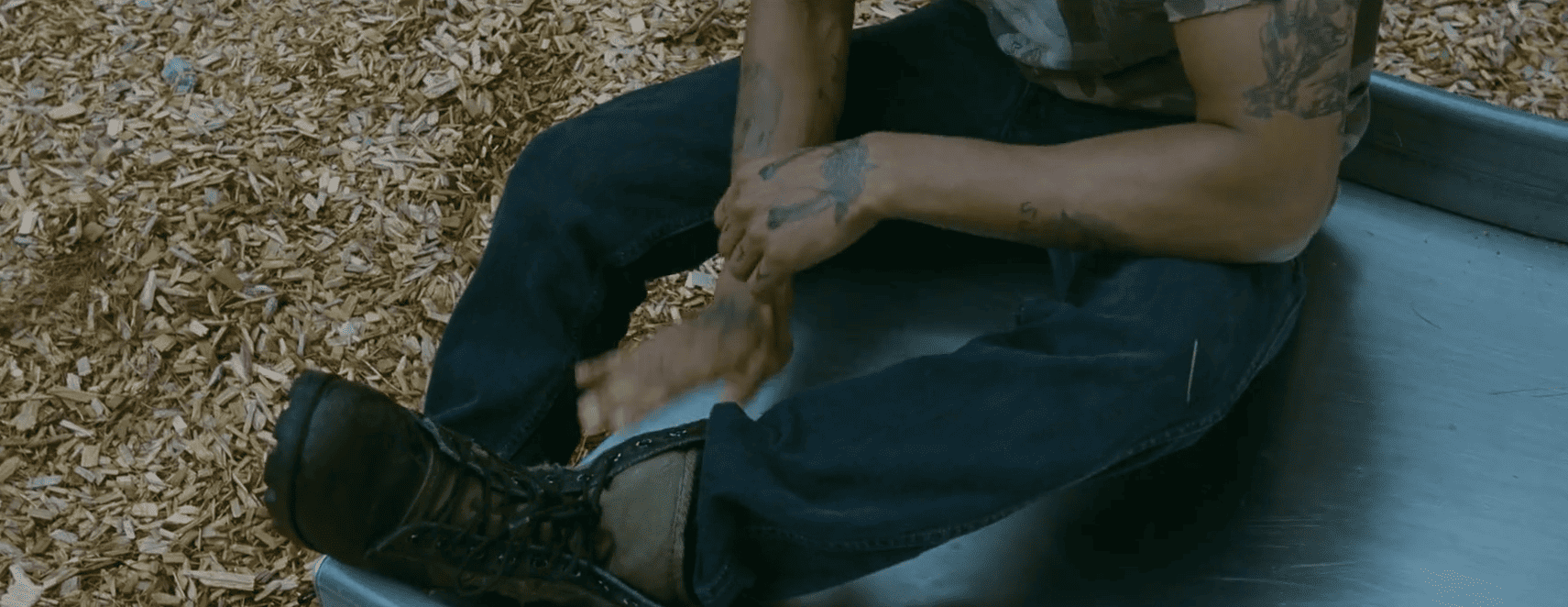 Les jambes et les bras d'un jeune homme musclé, à la peau mate. Il est en assis sur un toboggan metallique, lui-même posé sur des copeaux de sciure. Le jeune homme, aux bras tatoués, en jean et chaussures montantes, tape sur le toboggan de la main droite.