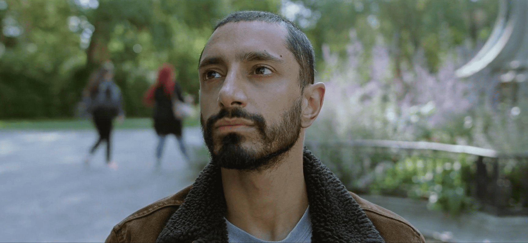 Un jeune homme brun, aux cheveux très courts, avec une courte barbe, en veste en jean marron doublée de laine de mouton, regarde vers le haut et la droite, dans l'allée d'un parc.