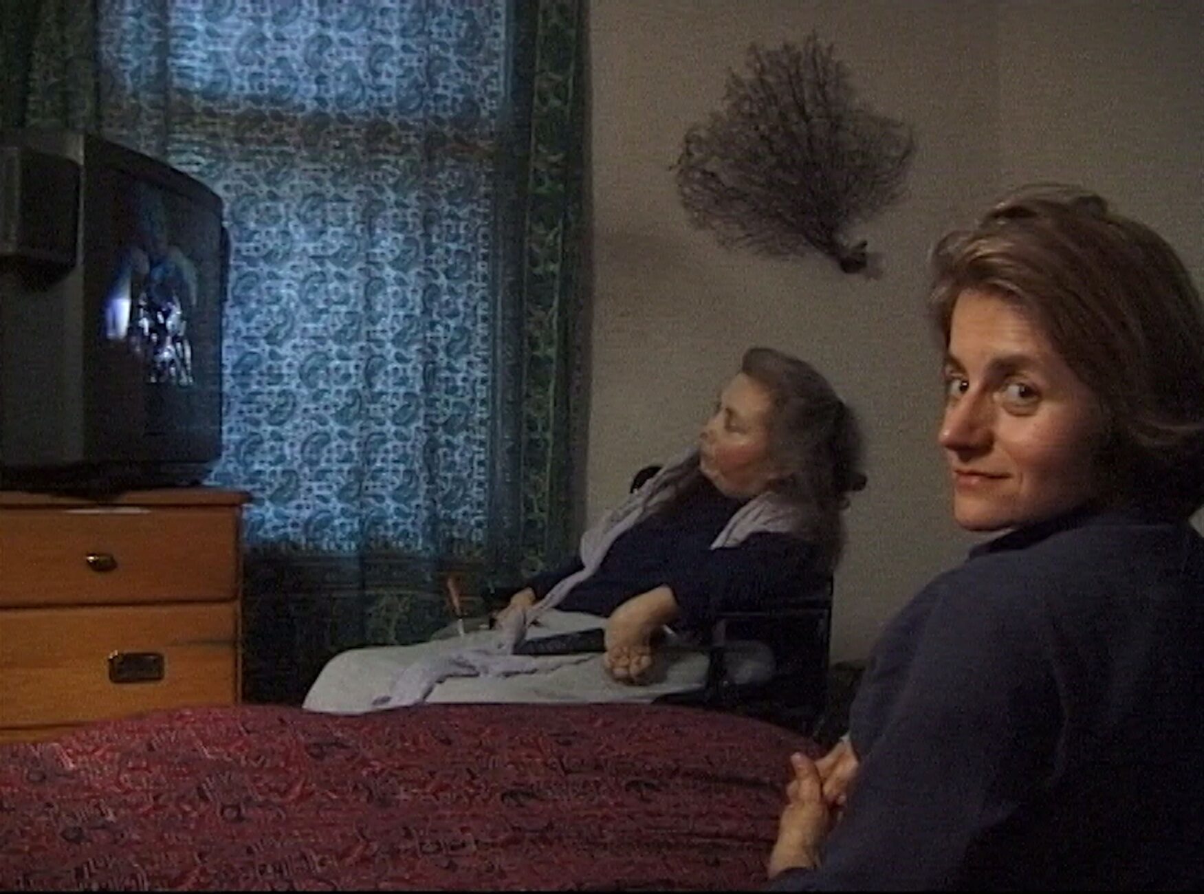Dans une chambre aux rideaux tirés, la poétesse Cheryl Marie Wade, cheveux gris et longs, est assise dans un fauteuil roulant et regarde un écran de télévision allumé devant elle. À sa droite, sur un lit à l'avant-plan, Diane Maroger, la trentaine et les cheveux mi-longs, tourne le visage vers nous.