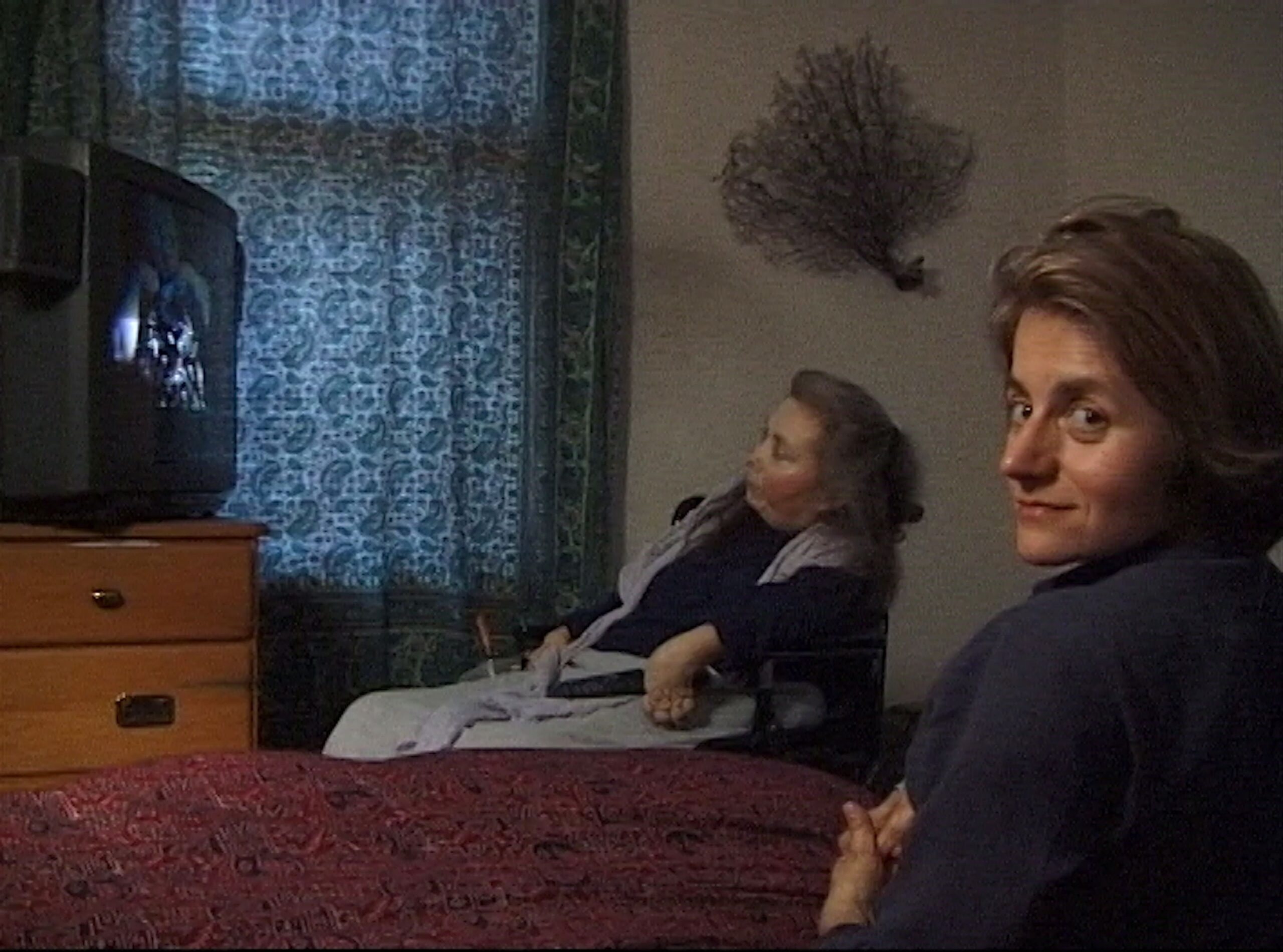 Dans une chambre aux rideaux tirés, la poétesse Cheryl Marie Wade, cheveux gris et longs, est assise dans un fauteuil roulant et regarde un écran de télévision allumé devant elle. À sa droite, sur un lit à l'avant-plan, Diane Maroger, la trentaine et les cheveux mi-longs, tourne le visage vers nous.
