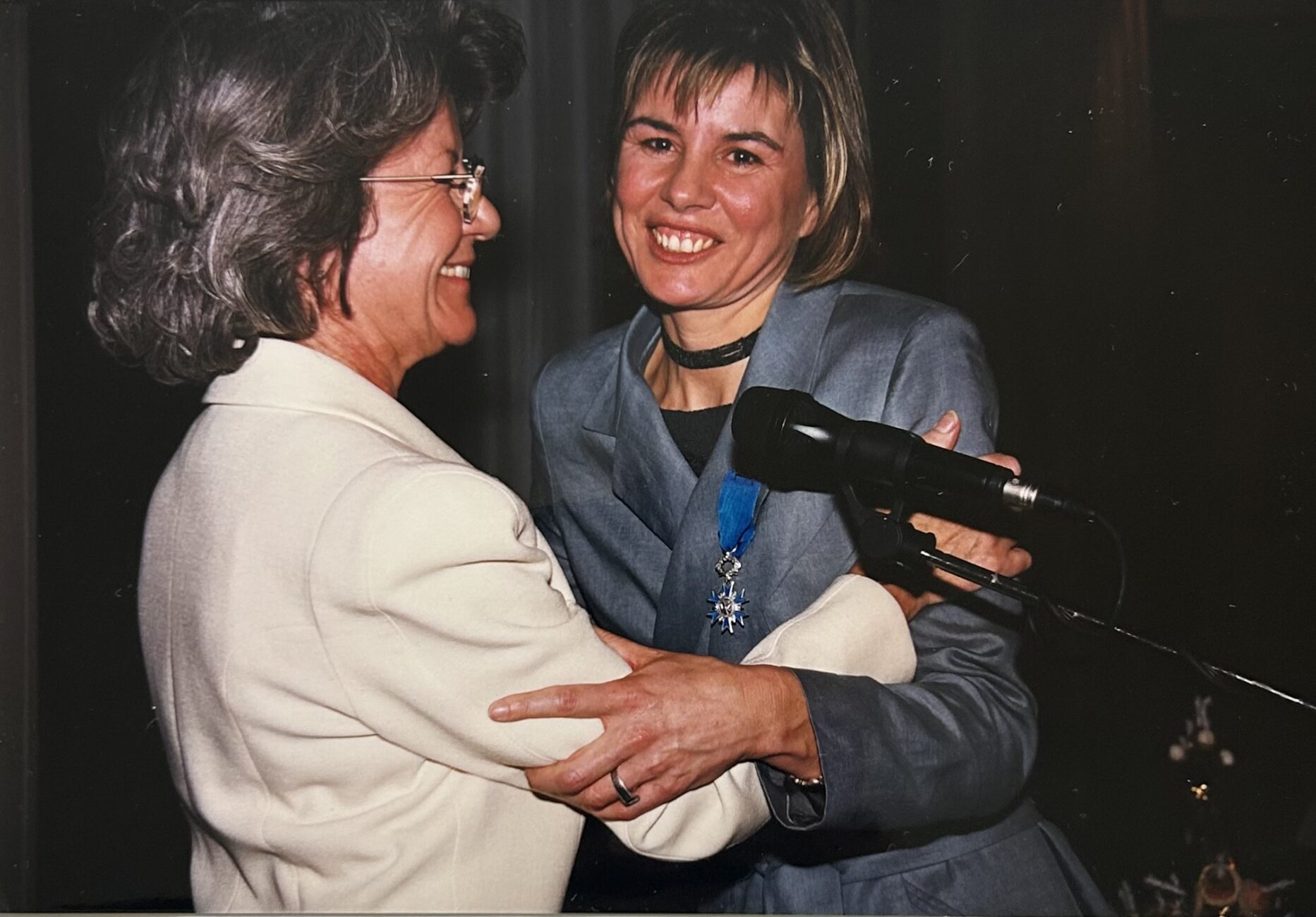 Une accolade entre Dominique Gillot, cheveux gris et tailleurs crème, et Marie-Thérèse L'Huillier en tailleur bleu.