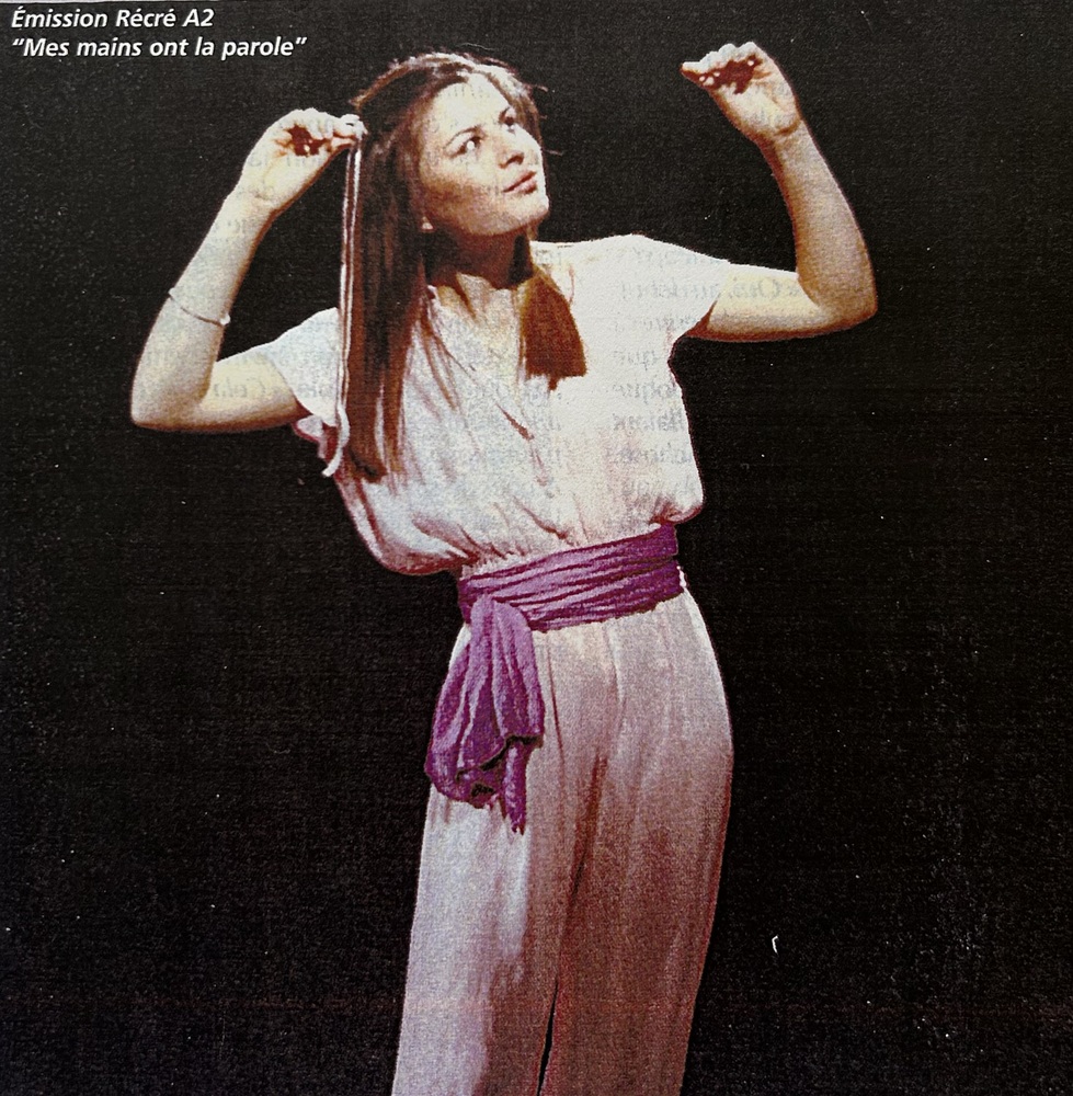 Marie-Thérèse L'Huillier en combinaison rose claire, ceinture en tissu rose. Les mains levées, elle regarde vers sa main gauche. En légende, sur la photo : "Emission Récré A2, Mes mains ont la parole".