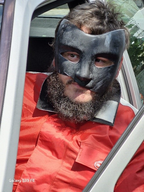 Un jeune homme est assis dans une voiture. Il porte une combinaison de travail rouge. Son visage est presque entièrement recouvert d’un masque noir, seuls apparaissent ses cheveux bruns au-dessus et sa bouche et sa barbe brune en-dessous. Il est encadré par la portière ouverte de la voiture et se penche légèrement dehors.