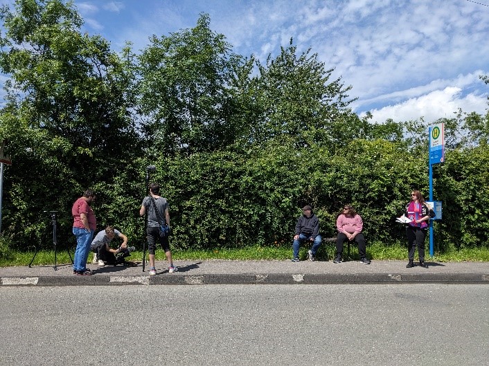 Six personnes se tiennent sur le trottoir, au bord d’une route secondaire. Un panneau et un banc indiquent la présence d’un arrêt de bus. Trois personnes s’affairent autour du matériel de tournage, deux jeunes hommes sont assis et une jeune femme, en maillot de foot avec une écharpe autour du cou, attend debout. La végétation est assez dense avec quelques arbres. Le ciel est bleu, parcouru de quelques nuages.