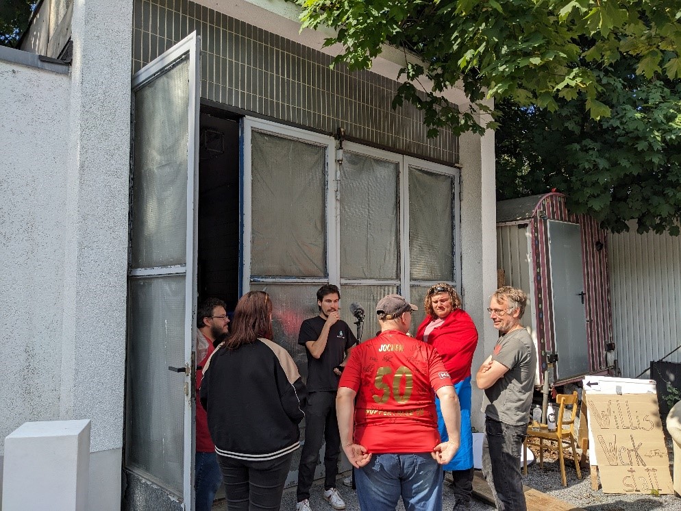Six personnes se tiennent debout en cercle devant la porte entrouverte d’un hangar. Les visages sont tournés vers l’homme le plus âgé. Un jeune homme en noir porte une perche avec un micro. Les quatre autres portent des vêtements aux couleurs du club de foot local. 